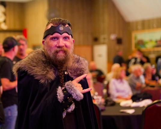 Man in fur-trimmed cloakand with a pointed leather band on his forehead makes sign of horns with crowded scene behind