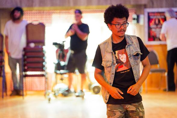 Young black man wearing anime shaded t-shirt and blue vest looks to the right with more people stuck to the wall behind him