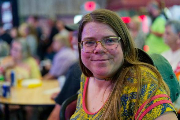 Smiling woman with long straight hair is wearing glasses ans has a hat slung over her back and a yellow and blue top