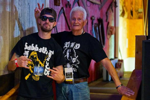Two men in black T-shirts:  left a younger man with baseball cap and Zakk Sabbath Tour 2025 and right an older many with a shirt that say DIO and white hair;  behind a wall with tools you might find in a country barn