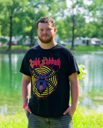 Man with short curly hair, short beard and zakk sabbath t-shirt with a purple spider on a yellow spiral with a pond behind with trees and campers past that