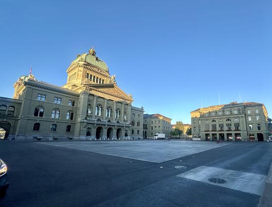 Bundesplatz in Bern, im Schatten. Die Kuppel des Bundeshauses ist von dem ersten Morgenlicht erhellt. Der Himmel ist strahlend blau.