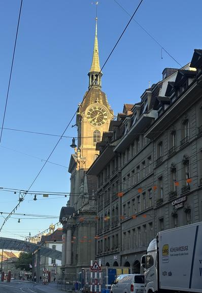 Der Kirchturm der Heiliggeist-Kirche in Bern unter strahlend blauem Himmel. Der Turm ist zur Hälfte ins frühe Morgenlicht getaucht. Das grosse Zifferblatt zeigt 6:40 Uhr.