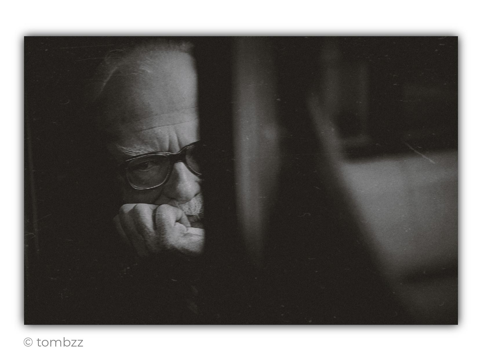 A black-and-white analog street photograph taken from outside, through the window of a bus. In the dim interior, an elderly man wearing large, dark-framed glasses sits with his face partly illuminated by soft light entering from outside. The light emphasizes the wrinkles on his forehead, his grey eyebrows, the outline of his nose, and his short facial hair. He rests his chin and mouth against a clenched hand, as though caught in a long, silent thought. His gaze seems directed beyond the bus, yet it feels turned inward rather than focused on anything outside.
A vertical window pillar cuts through the centre of the frame and conceals part of his face, strengthening the sense of separation and of witnessing a brief, private moment. The right side of the photograph remains blurred and almost abstract, with a pale rectangular area and dark bands from the bus interior. Fine scratches, dust, and marks are visible on the glass, reinforcing the raw analog character of the image. The whole scene feels quiet, intimate, and deeply contemplative.