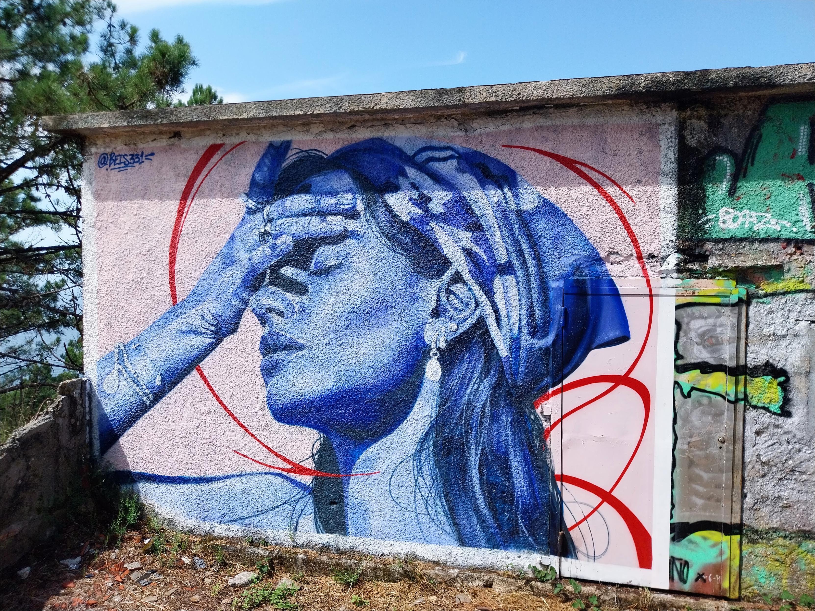 Gemälde einer jungen Frau an der Wand eines Gebäudes. Man sieht den Kopf in Seitenansicht, umschlungen von einem Kopftuch, unter dem lange Haare über die Schulter fallen, sie hält die Augen geschlossen, mit der Hand fasst sie sich an die Stirn. Sie trägt Schmuck an Ohren, Arm und Fingern. Das Gemälde ist ganz in Blautönen gehalten.