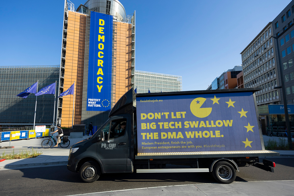 A photo of the European Parliament with a blue banner stating "DEMOCRACY. Protect what matters". In front, on the street, a van with a banner "DON'T LET BIG TECH SWALLOW THE DMA WHOLE" and pacman eating the EU member stars