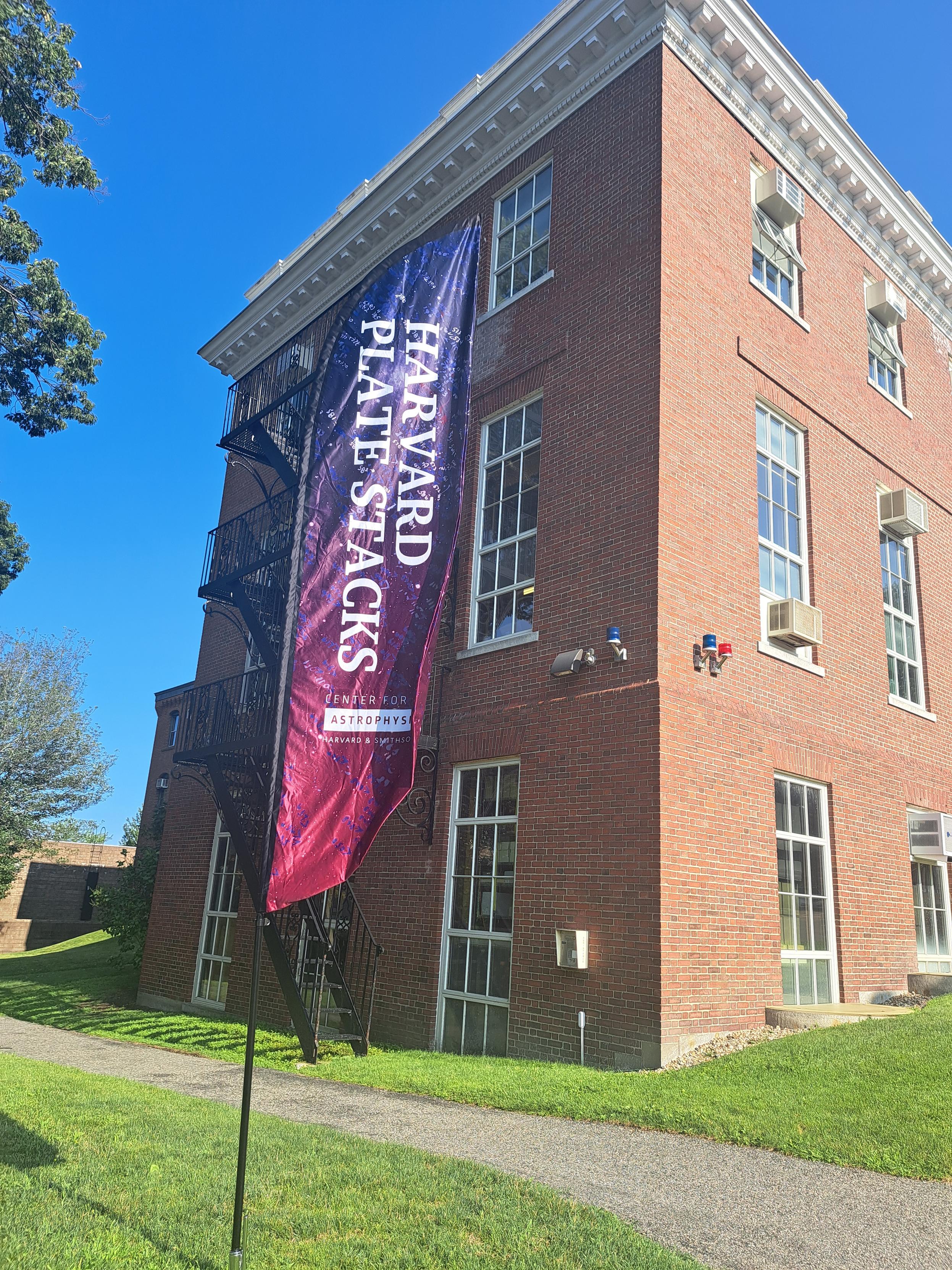 A brick building in front of which is a purple-blueish flag/sign saying "Harvard Plate Stacks" and the "Center for Astrophysics | Harvard & Smithsonian" logo.

The brick building (labelled A in CfA parlance) is the historical home of said plate stacks.