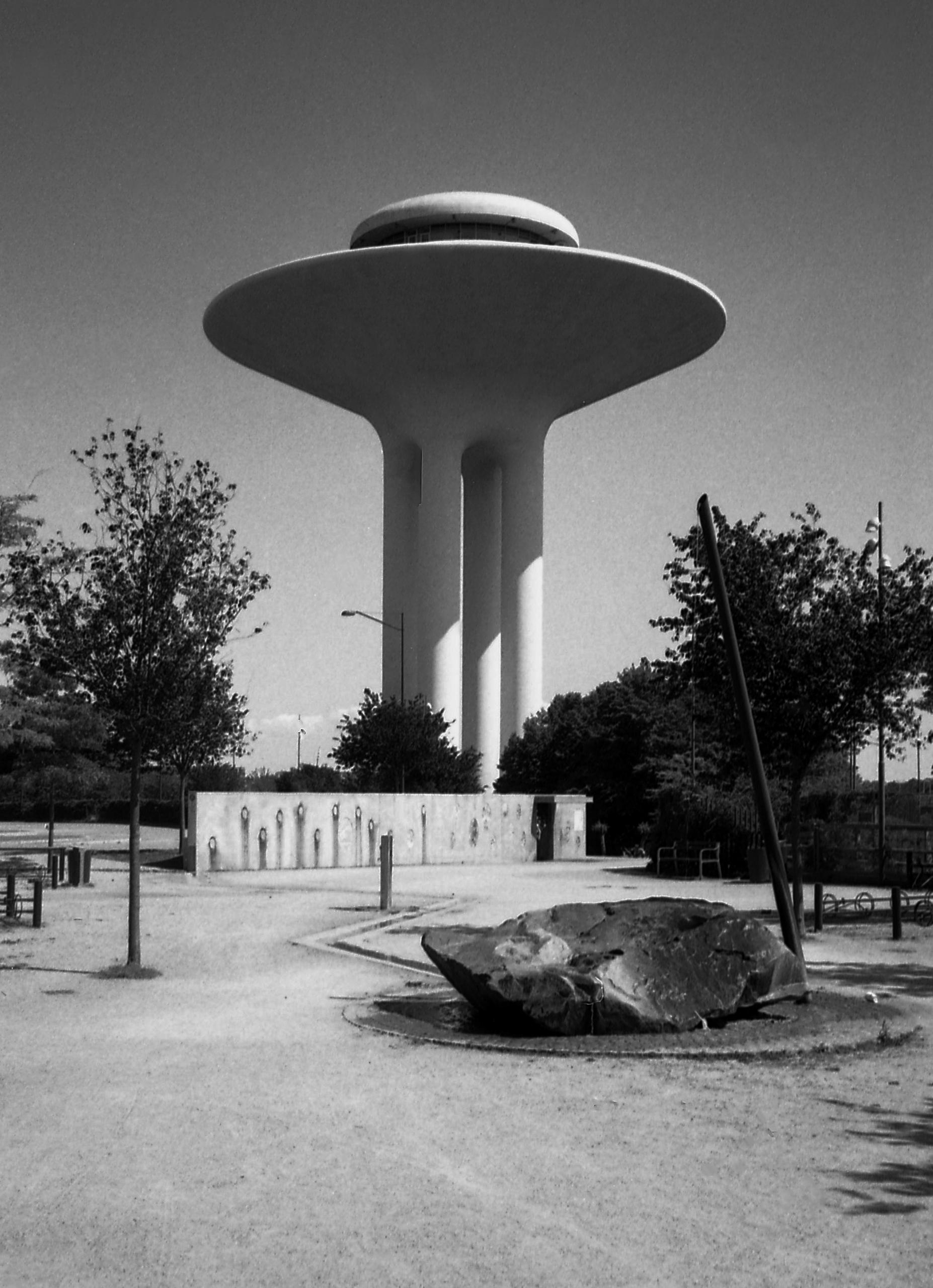 Ein Wasserturm, der aussieht wie ein UFO sud Stelzen. Auf dem Platz vor dem Turm liegt ein Findling und stehen ein paar Bäume