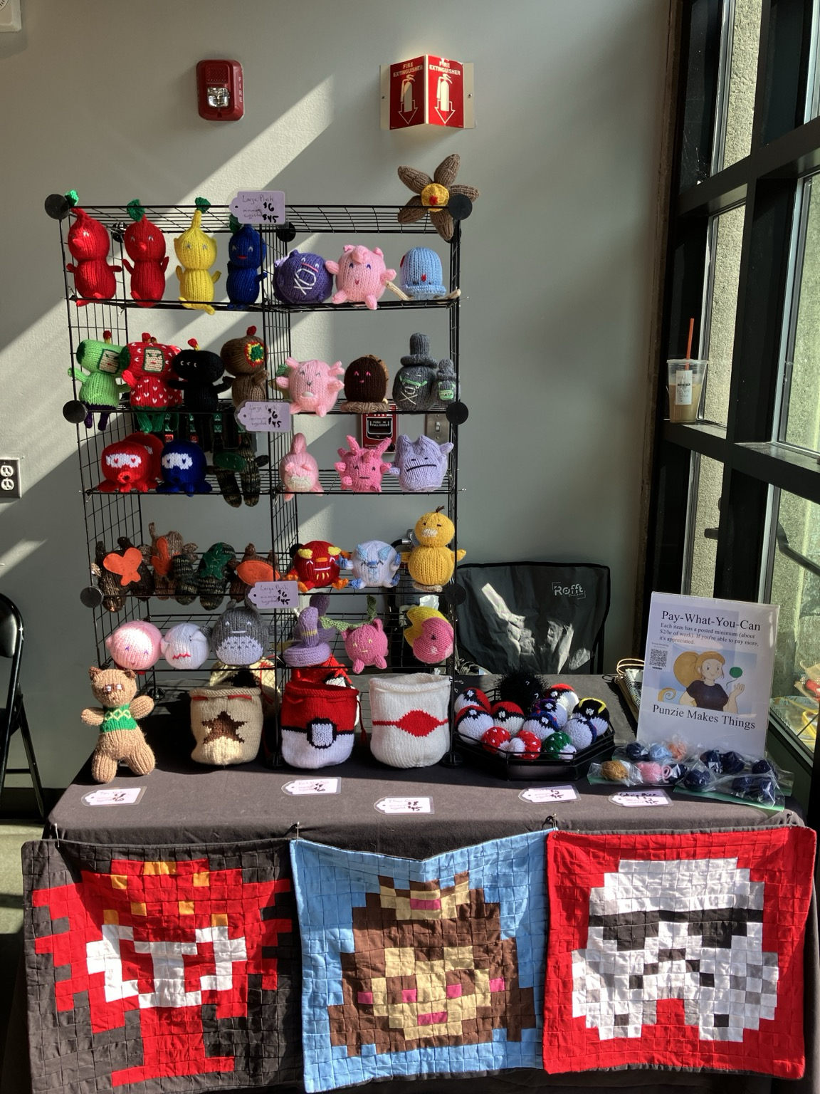 Vendor table with multiple video game-themed knit plush like pokemon, koroks, Tom Nook, katamari, and pikmin. There are also some knit dice bags. Across the bottom front of the table are 3 pillow covers with 8-bit images of an octorok, princess peach, and a storm trooper
