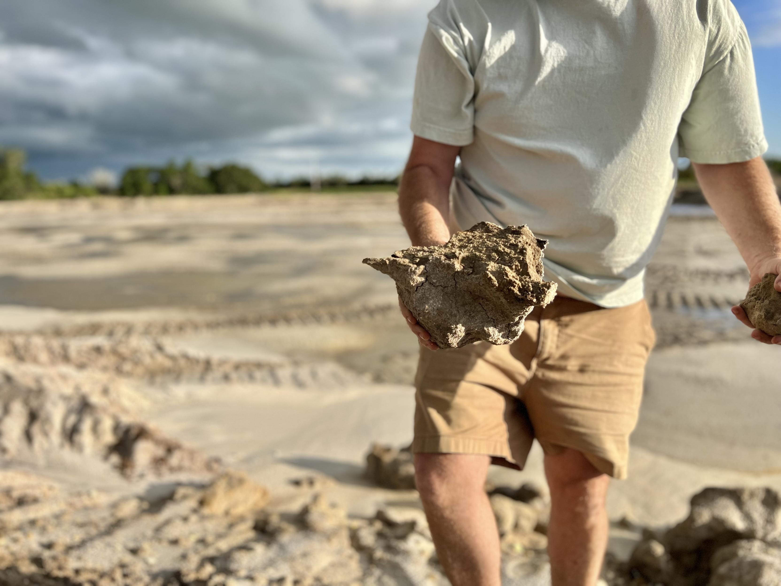 A photo of my husband holding a large hunk of sandy clay. His head is cut off so you can just see his body from his shoulders to his calves. He’s wearing a light blue t-shirt and tan shorts. He’s standing on a sandy gray construction site. There is a line of trees and a cloudy sky far off in the background.