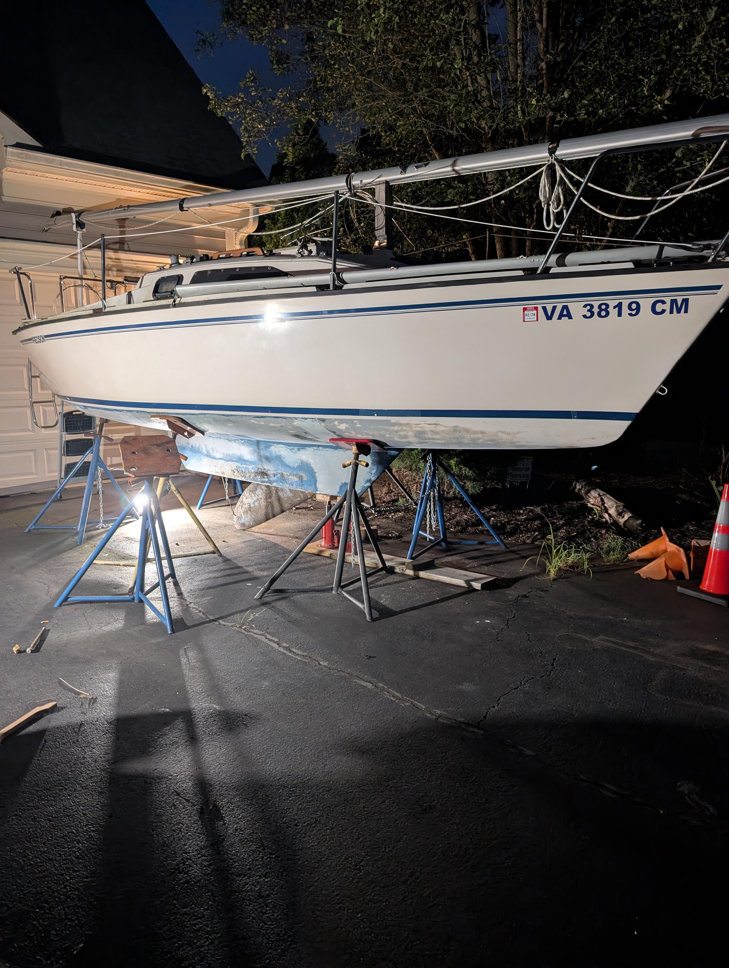 A Precision 21 sailboat sits on six triangular based boat metal pipe boat stands, placed at the stern, middle and bow of the boat. The mast is down, laying down the middle of the length of the boat. The swing keel has come out of the keel housing and hangs at an angle from the keel of the boat onto the asphalt driveway.  It is night, so there are small Milwaukee work lights illuminating the scene