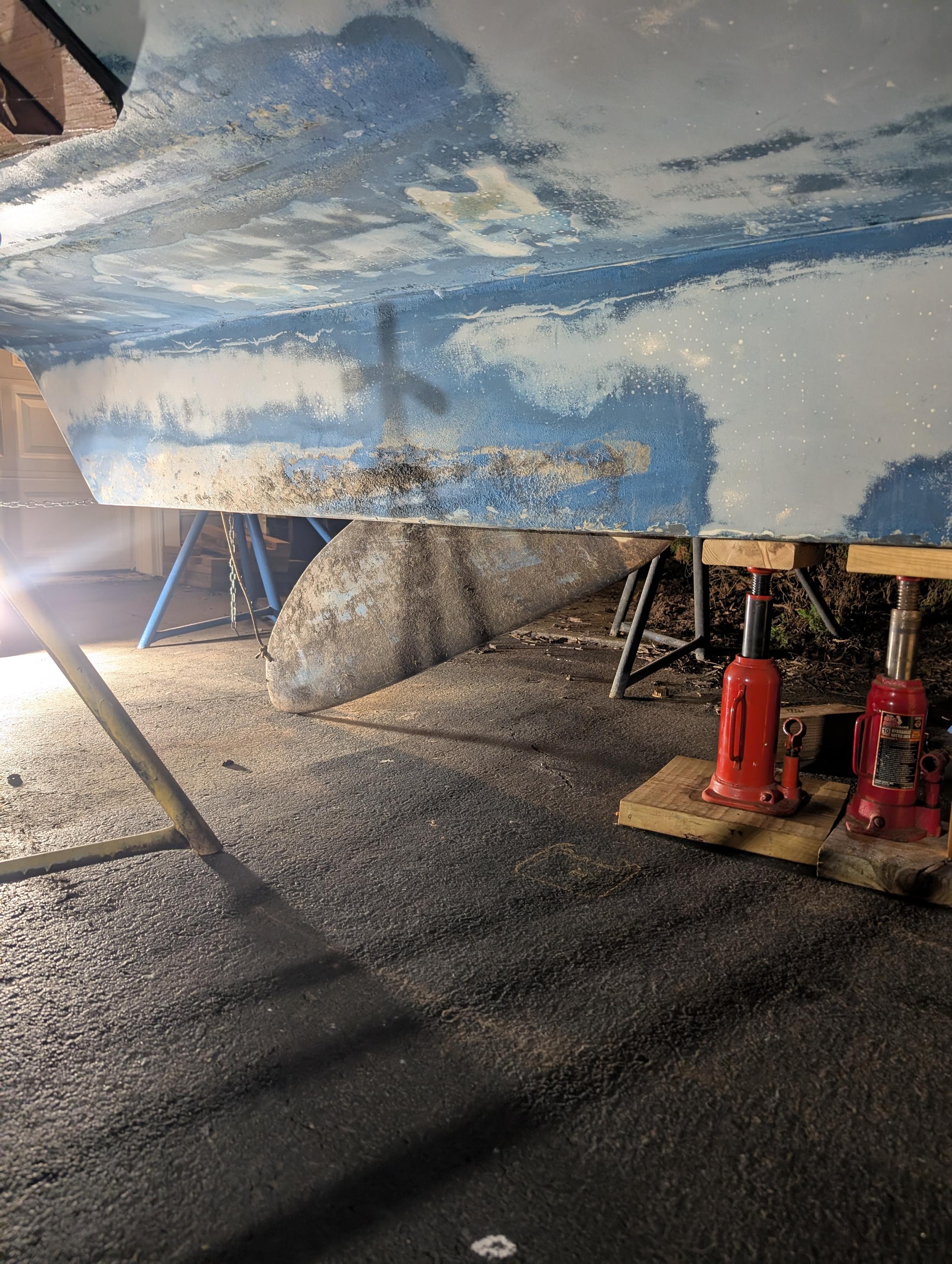 This is a close up of the swing keel that dropped from the keel housing. It is covered in grungy, dried brown algae growth. The keel housing base color is light taupe-gray, but there is a medium marine blue paint that remains unsanded on the face of the keel housing. There are two red and black bottle jacks holding up the front of the keel.of the boat, where the 1500lb lead keel weight is located. Everything is sitting on an asphalt driveway