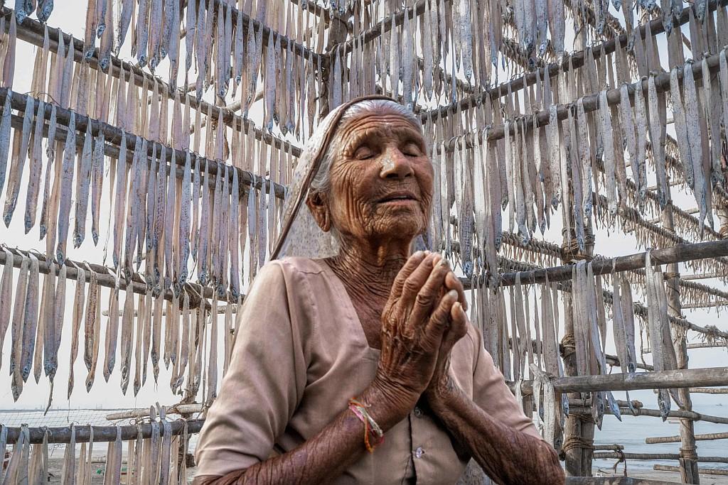 In a photograph by Nicolò Brugnara, an elder of the Indigenous Koli community in Mumbai stands with eyes closed and hands clasped in prayer. She is positioned inside a tall bamboo drying frame where hundreds of slender, silver Bombay duck (Harpadon nehereus) are suspended vertically to cure in the coastal air. Through the open lattice of the wooden structure, the coastal shoreline and pale sky are visible in the background.