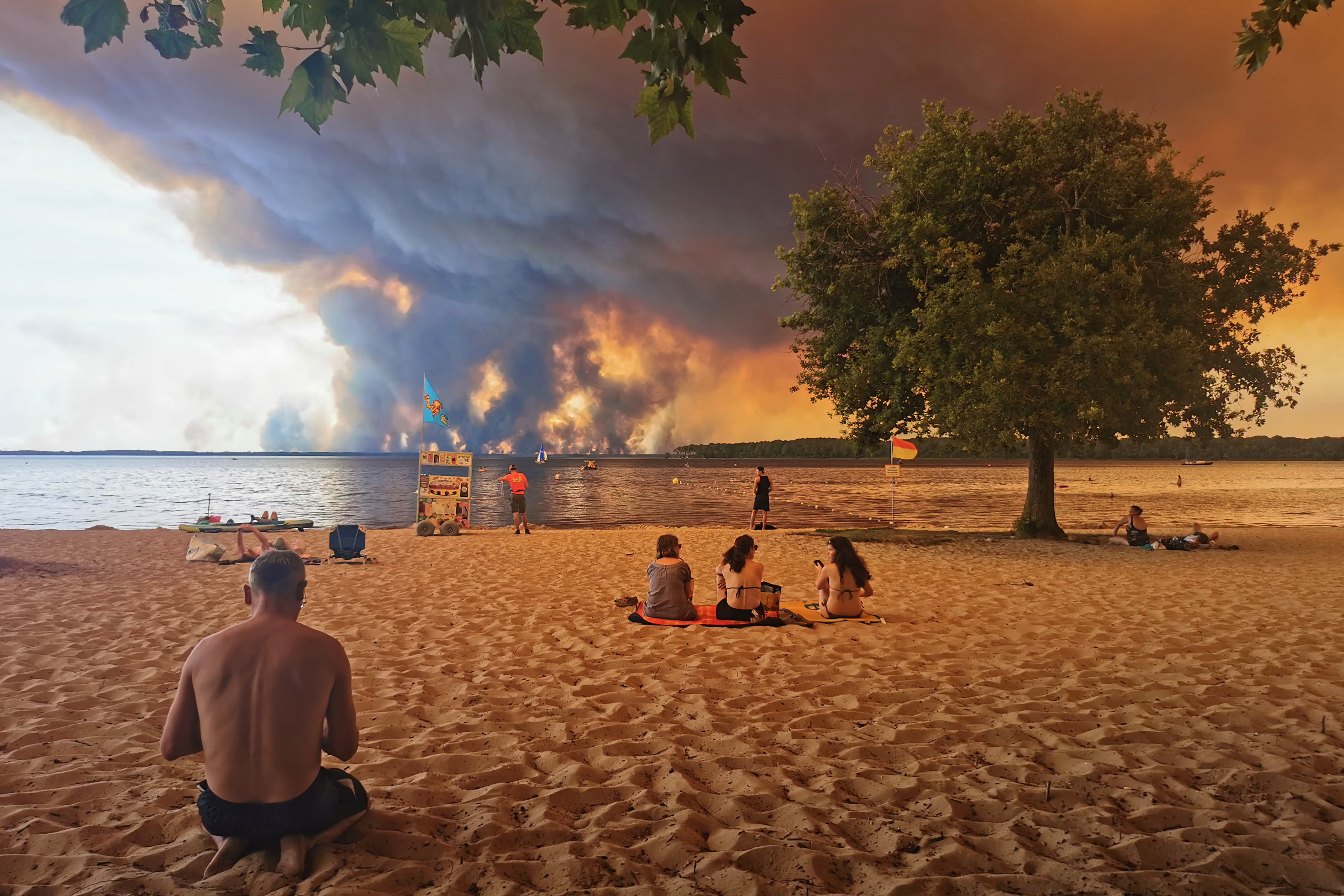 People sit on the beach of Moutchic as smoke rises into the sky from a wildfire. 