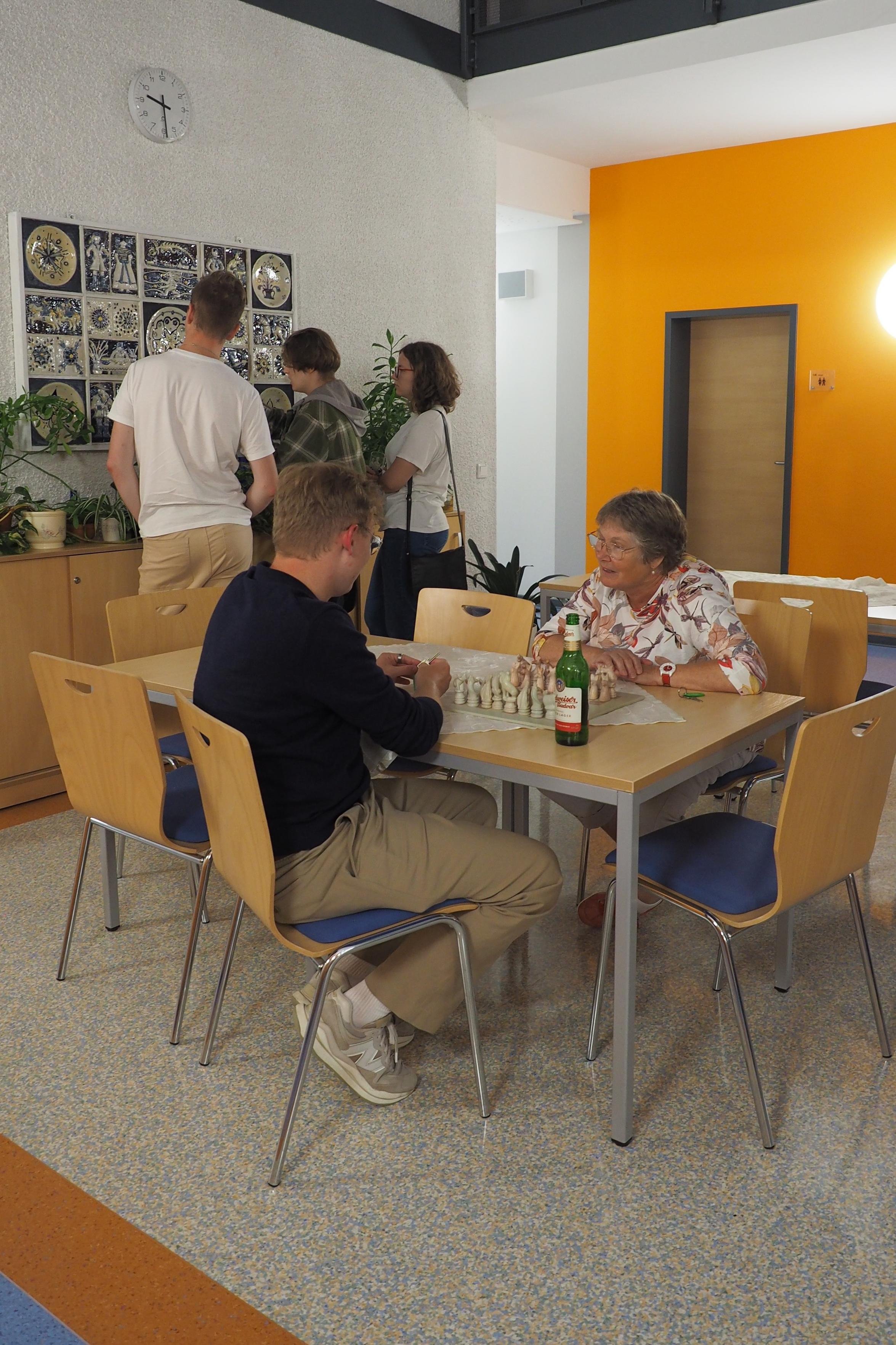 Das Bild zeigt zwei Menschen an einem Tisch, die gerade Schach miteinander spielen. Im Hintergrund schauen sich drei weitere Menschen auf Kunst an der Wand.