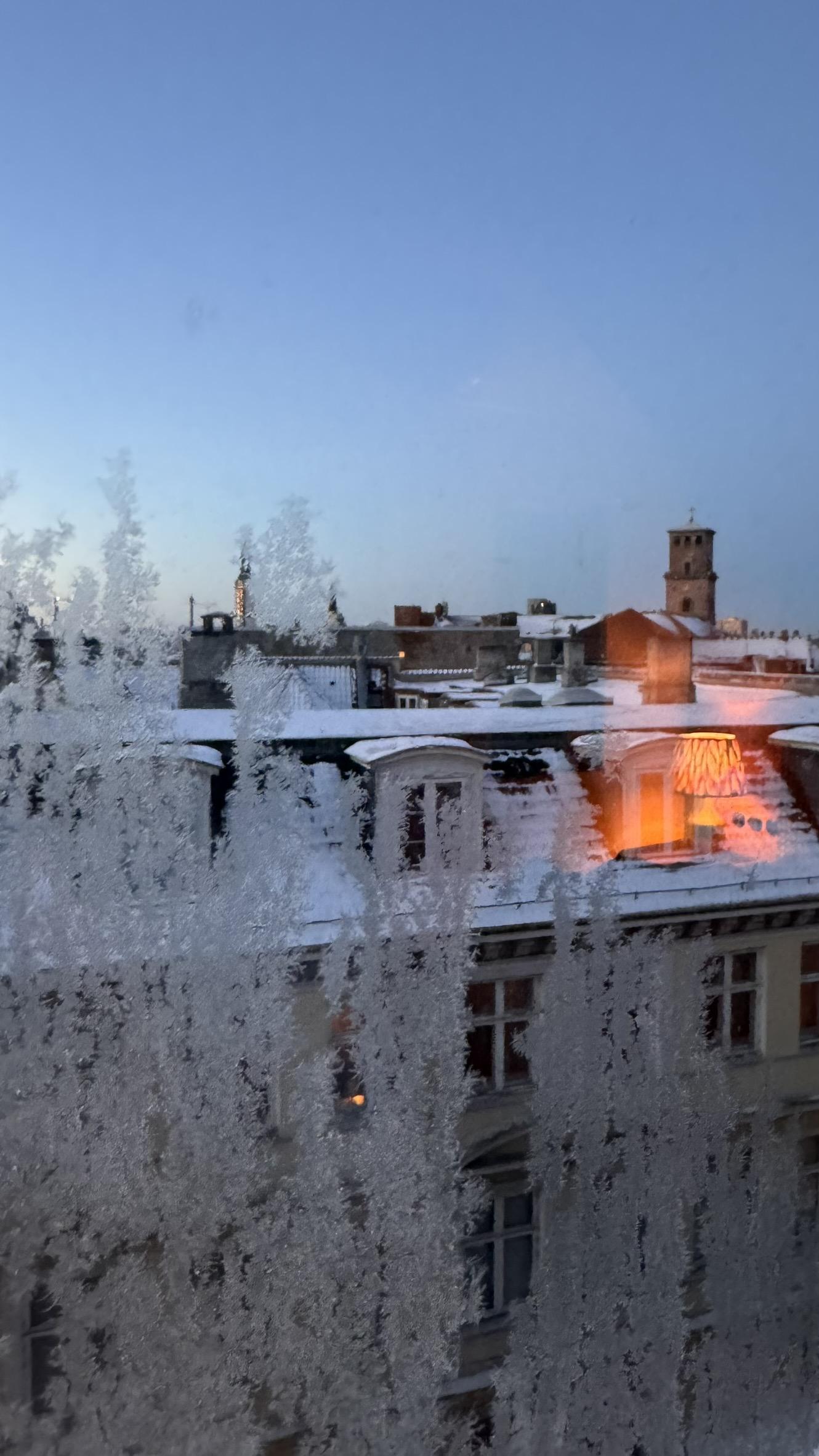 Kold, skyfri januar-morgen, snedækkede Københavnske hustage med Vor Frue Kirke i baggrunden. Taget gennem delvist tilfrosset vindue fra et værelse, højt oppe i 25hours Hotel Indre By. Som varm kontrast til de iskolde farver udenfor ses sengelampens varme genskær i vinduet.