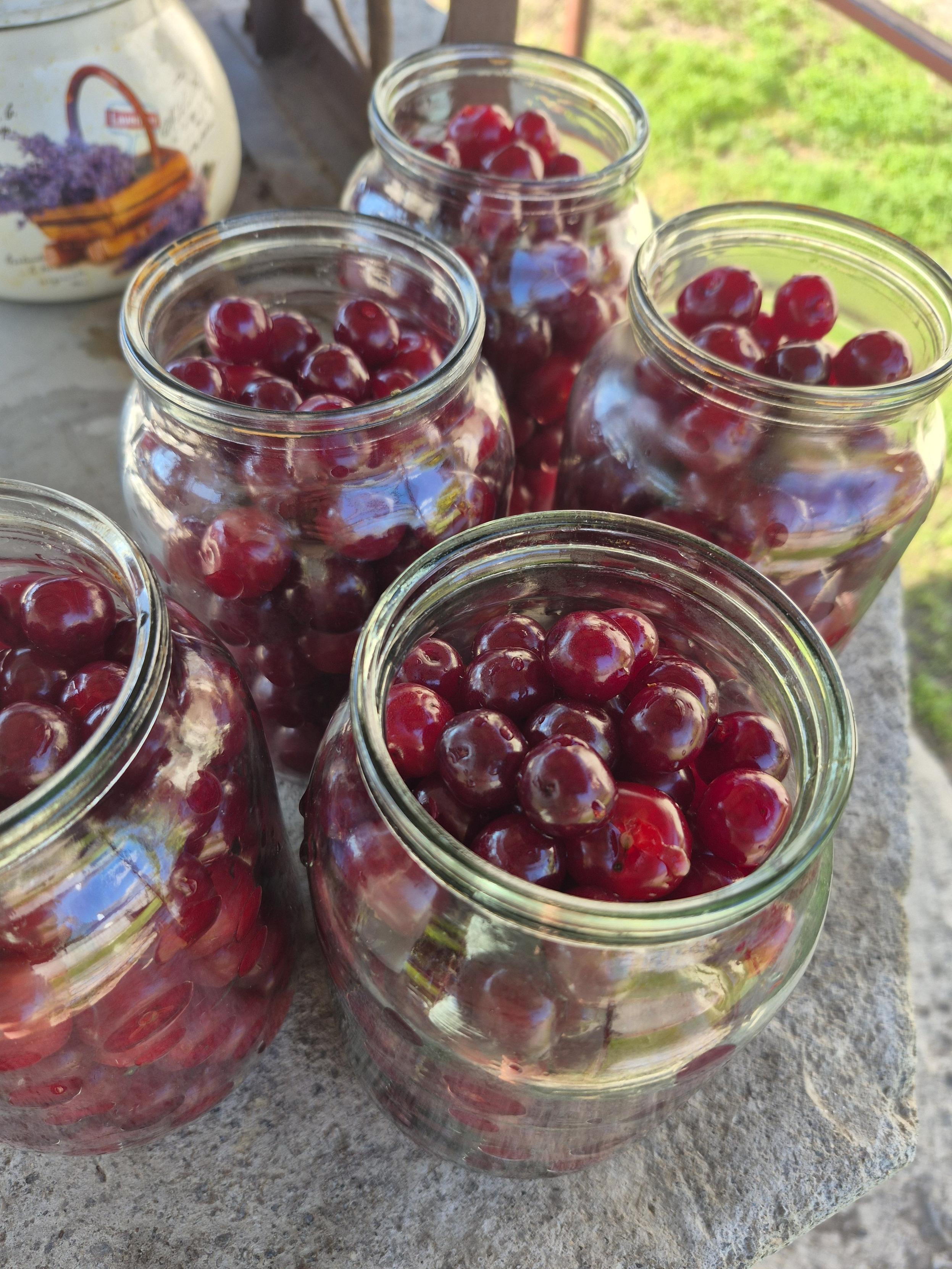 Several glass jars filled with fresh, dark red cherries, standing on a stone surface outdoors