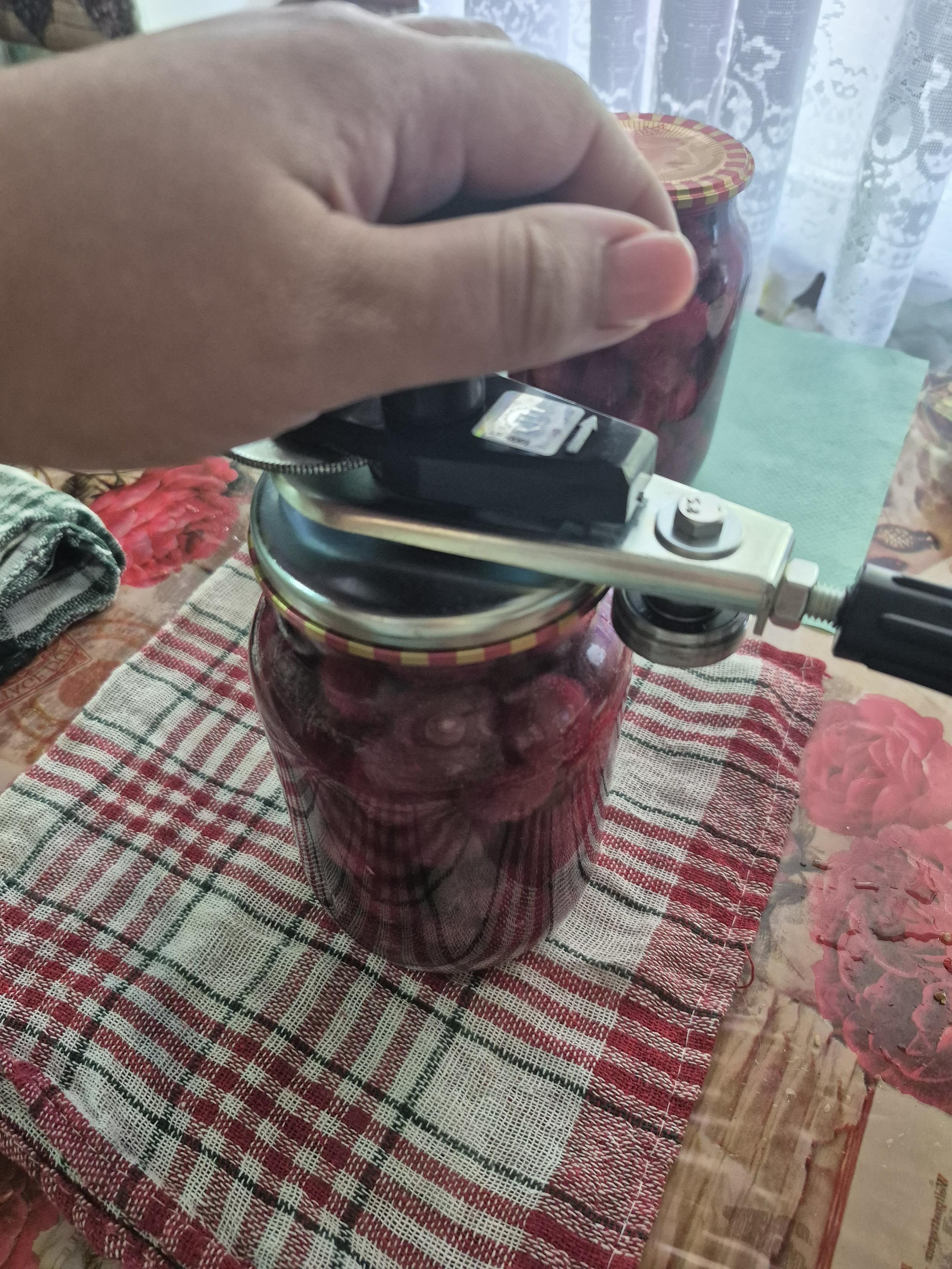 A hand using a manual can sealing machine to close a glass jar full of cherries on a plaid towel.