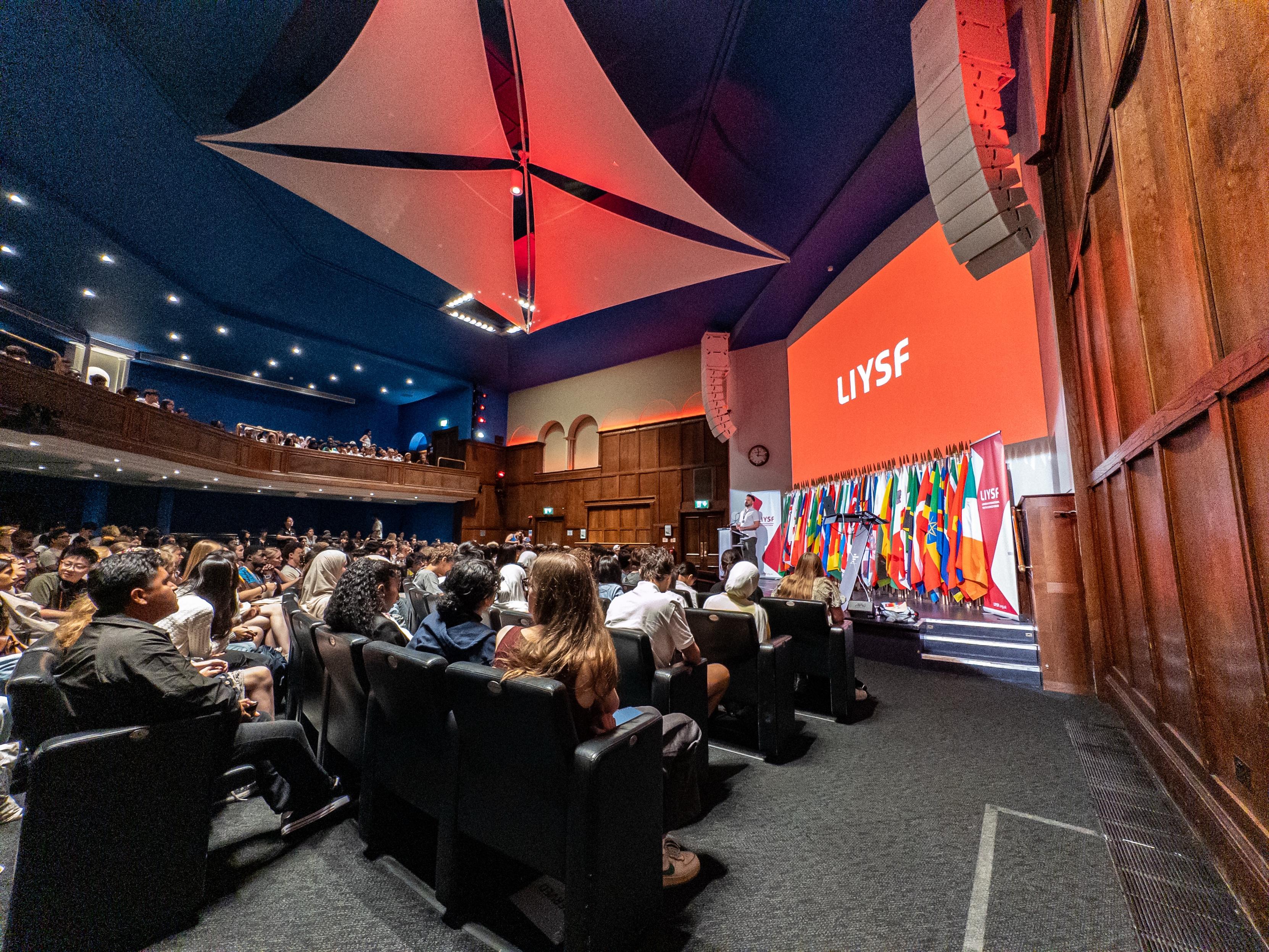 Another view into the auditorium, showing the podium with many flags and a large red screen with LIYSF written in white.