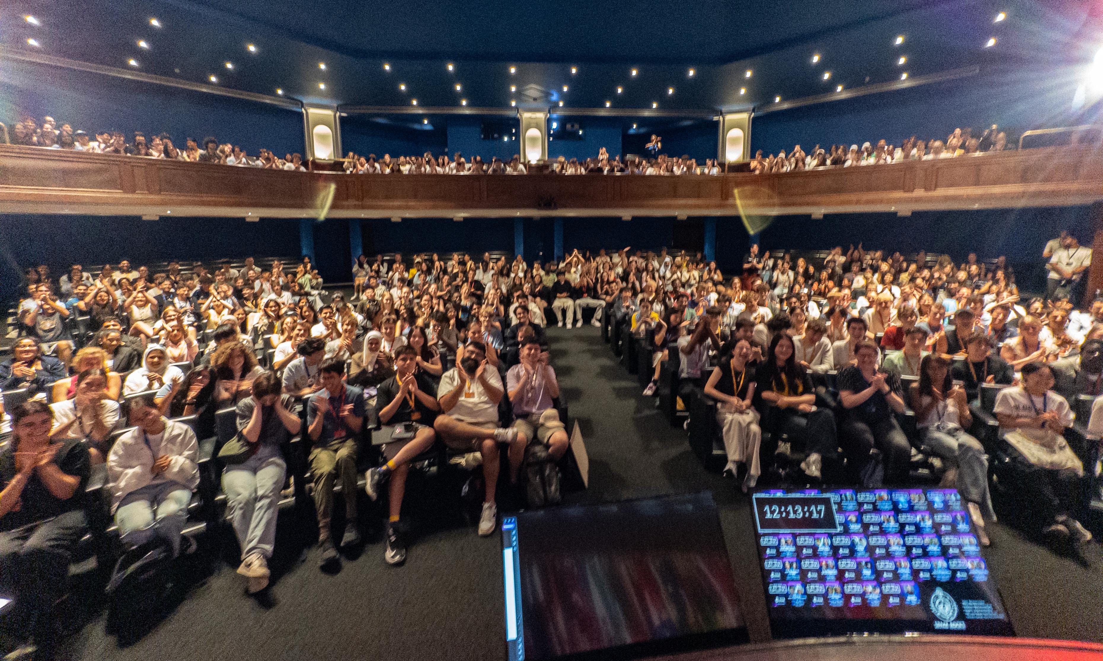 A large auditorium with many young people clapping, and some large computer monitor screens in the front.