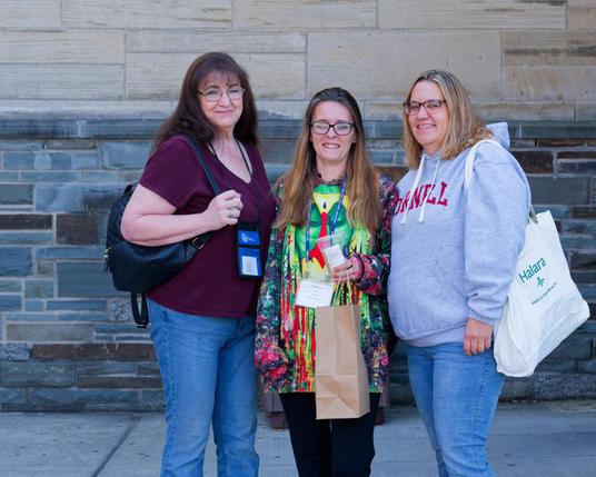 Three woman in front of a rock wall; tallest on the left with a dark purple blouse and black bag, shorted in the middle with a colorful top and brown bag and medium on the right with a cornell sweatshirt