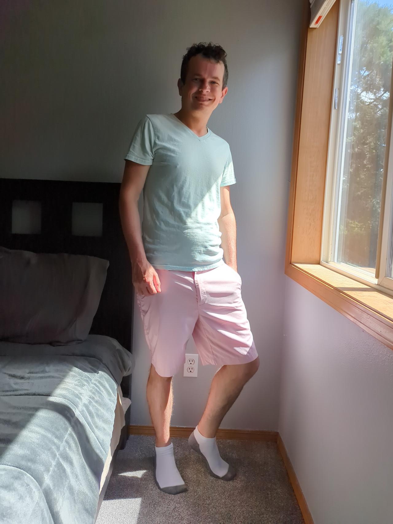 A fully clothed, full body photo of me standing next to the window in a bedroom. My face is grinning at the camera and I am wearing a light blue t-shirt, pink shorts, and white and grey ankle socks.