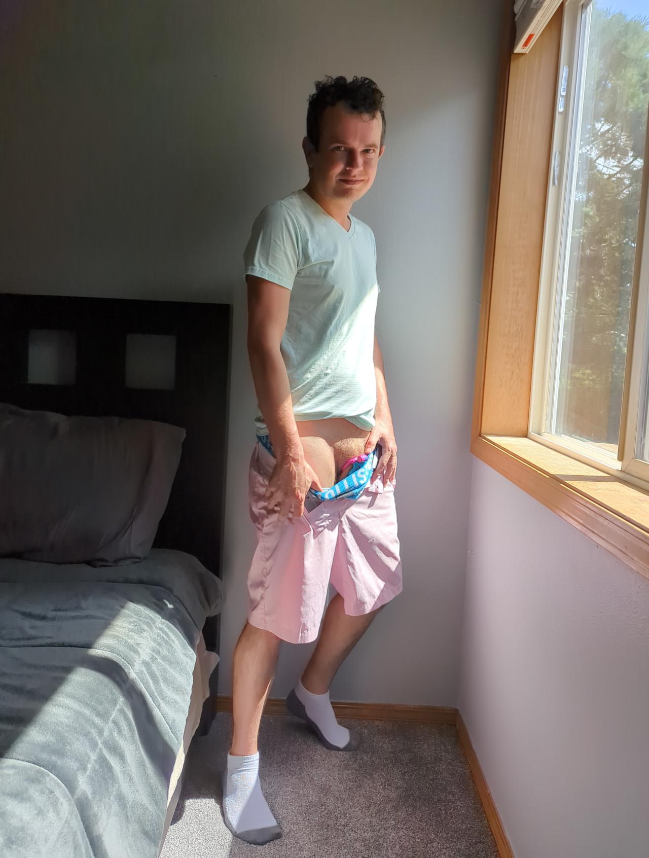 Now, my face looks at the camera with a sly, closed mouth smirk as my right hand pulls down the waistband of my blue underwear, revealing a slight teasing look at my junk and part of the chastity cage locked around my penis, and my pubic hair just above my junk glistens in the sunlight. The cage has a pink ring that is looping around under my balls and a blue cage which can only barely be seen.