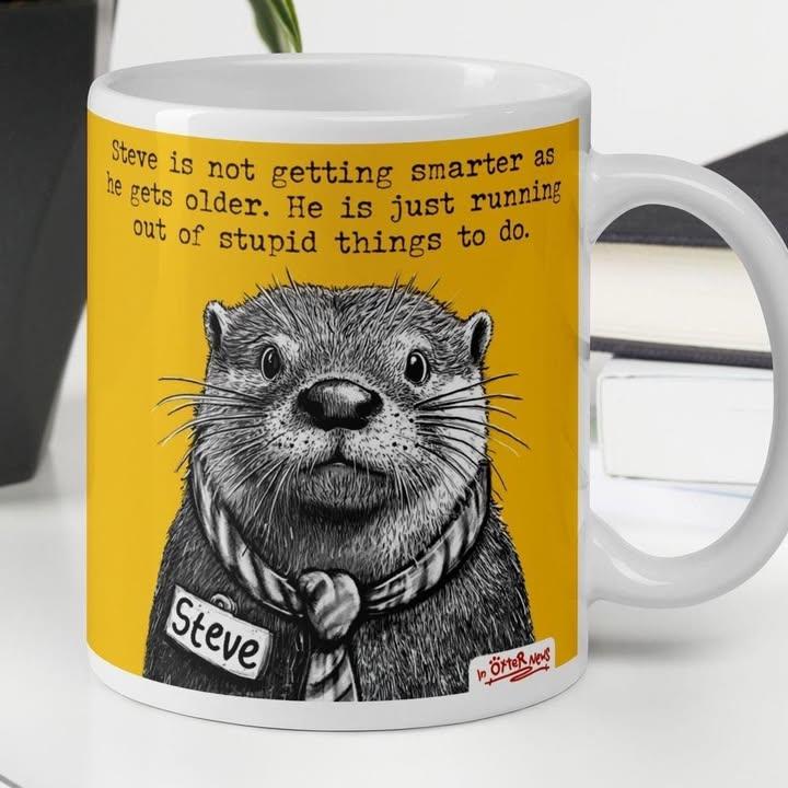 A white ceramic mug sitting on a desk displaying a whimsical black-and-white sketch of an otter in a scarf against a vibrant background.