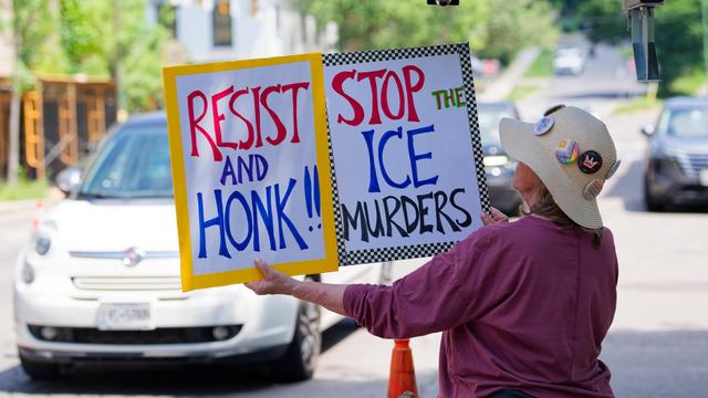 Woman with a hat covered with buttons holds signs that say RESIST and HONK and STOP THE ICE MURDERS seen from behind as traffic approaches behind