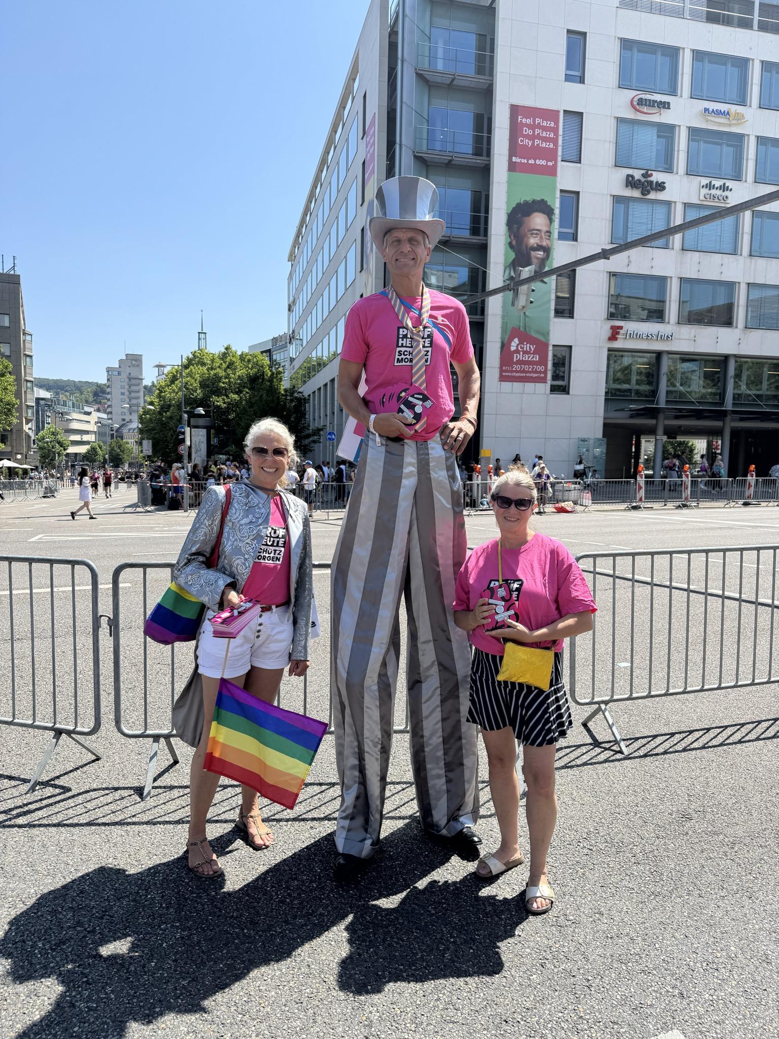 Zwei Damen mit PRÜF-T-Shirts stehen neben einem Stelzenläufer mit Zylinder und Prüf-T-Shirt (diese sind rosa und haben das PRÜF-Logo vorne drauf (siehe Bild links)