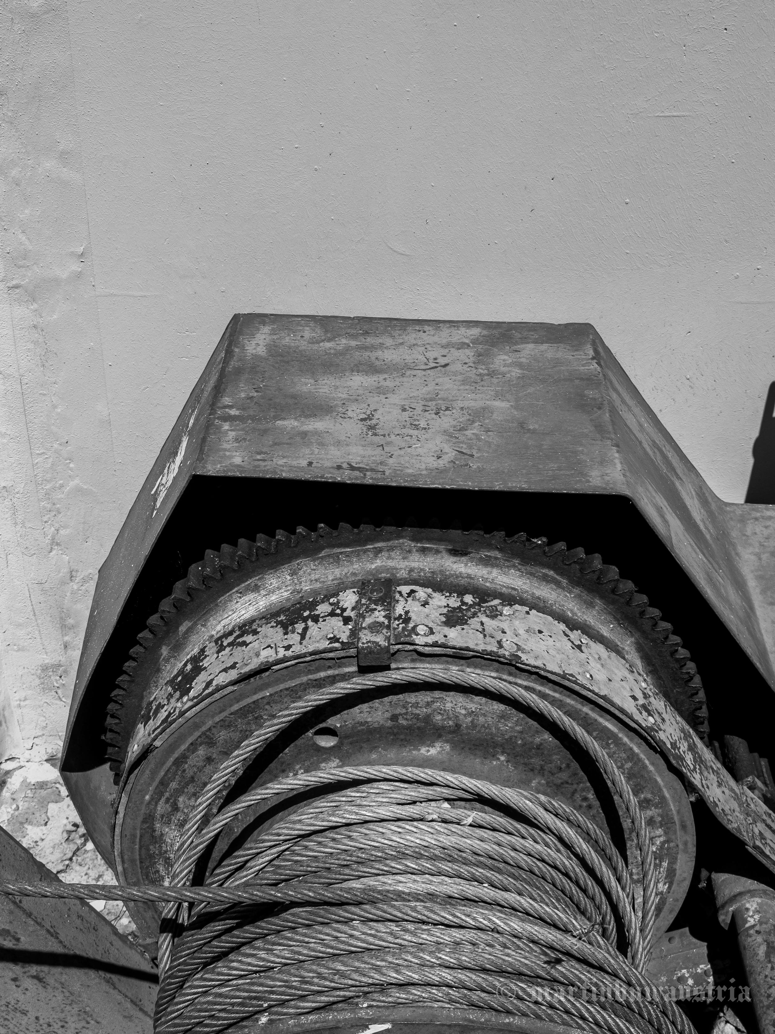 A black and white photo of a historic electric winch featuring a large cable drum, a thick steel cable, a protective cover, and visible traces of rust and aging.