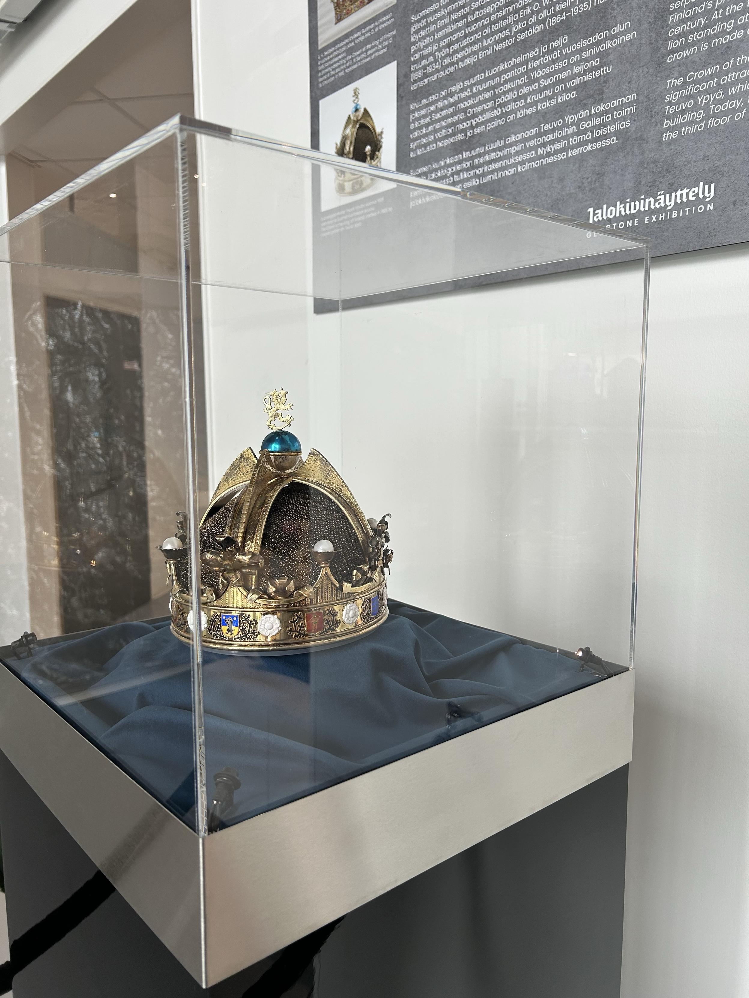 A kings crown in gold with stones in a glass display. On the wall is some text about the crown.