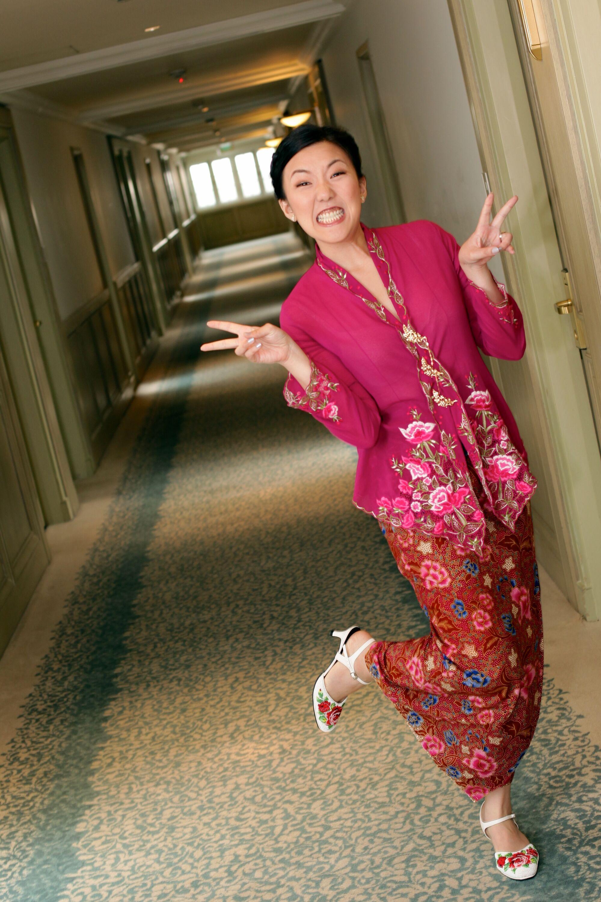 Joy in a traditional Peranakan Nyonya outfit at her wedding in Singapore. She's making a silly face and flashing two peace signs with her foot up.