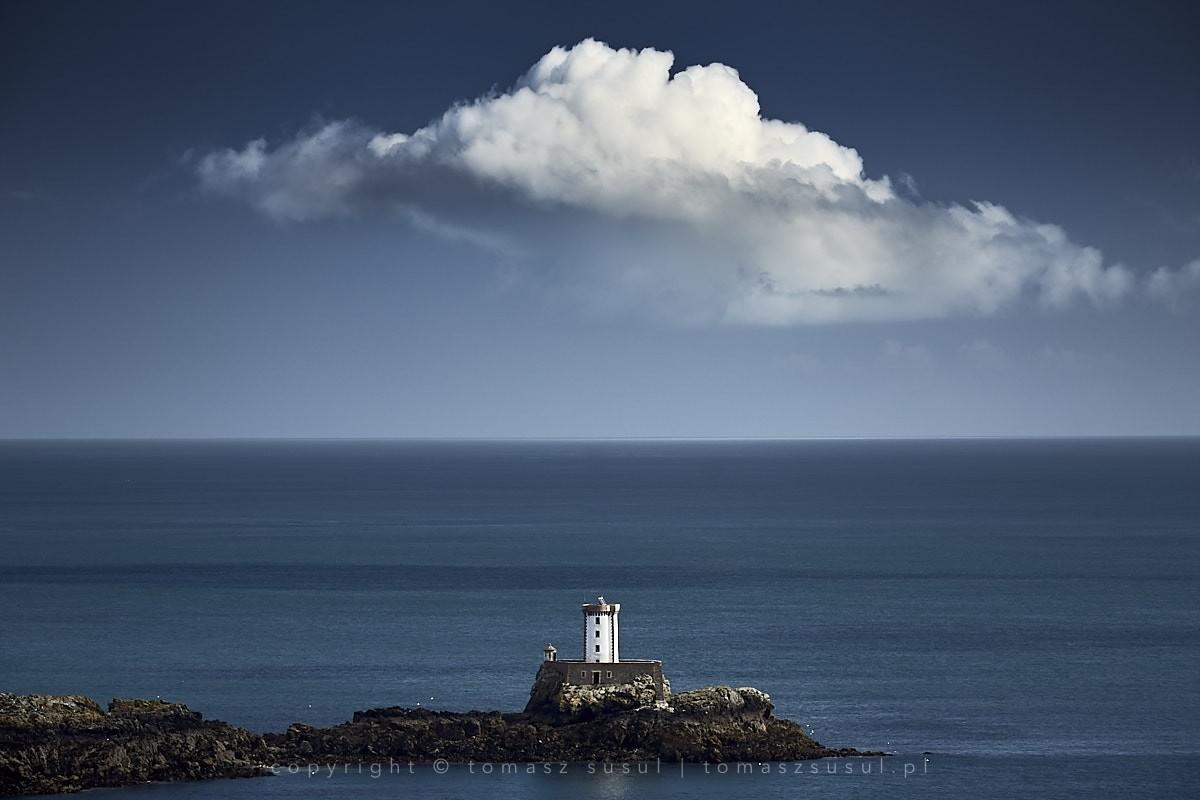 A compact white lighthouse stands perched upon a rugged rock formation amidst the sea. The surrounding water stretches toward the horizon, blending into deep blue tones of the sky in the distance. Above it, a massive, sculpted cloud towers over the sea, its immense size dwarfing the small lighthouse below.