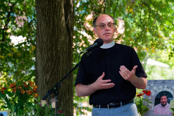 Minister with dark shirt and white collar speaks into microphone with his hands held up, the portrait of a people killed by ICE at the lower right and a tree trunk and spreading leaves on his left
