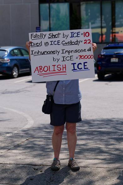 In a bank parking lot a person holds a sign in from of the their face that says "Fatally Shot by ICE = 10, Died in ICE Custody=22, Inumanhely Imprisoned by ICE=70,000. ABOLISH ICE"