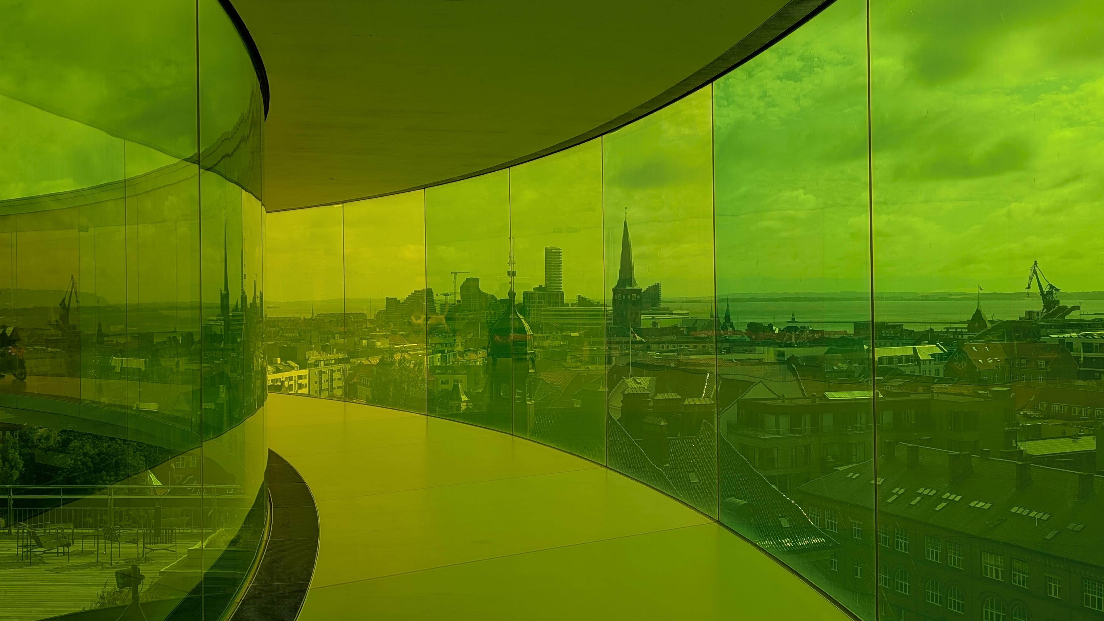 The skyline of Aarhus seen through yellow and green-coloured glass being part of a round skywalk, a church in front, modern skyscrapers and the sea in the back