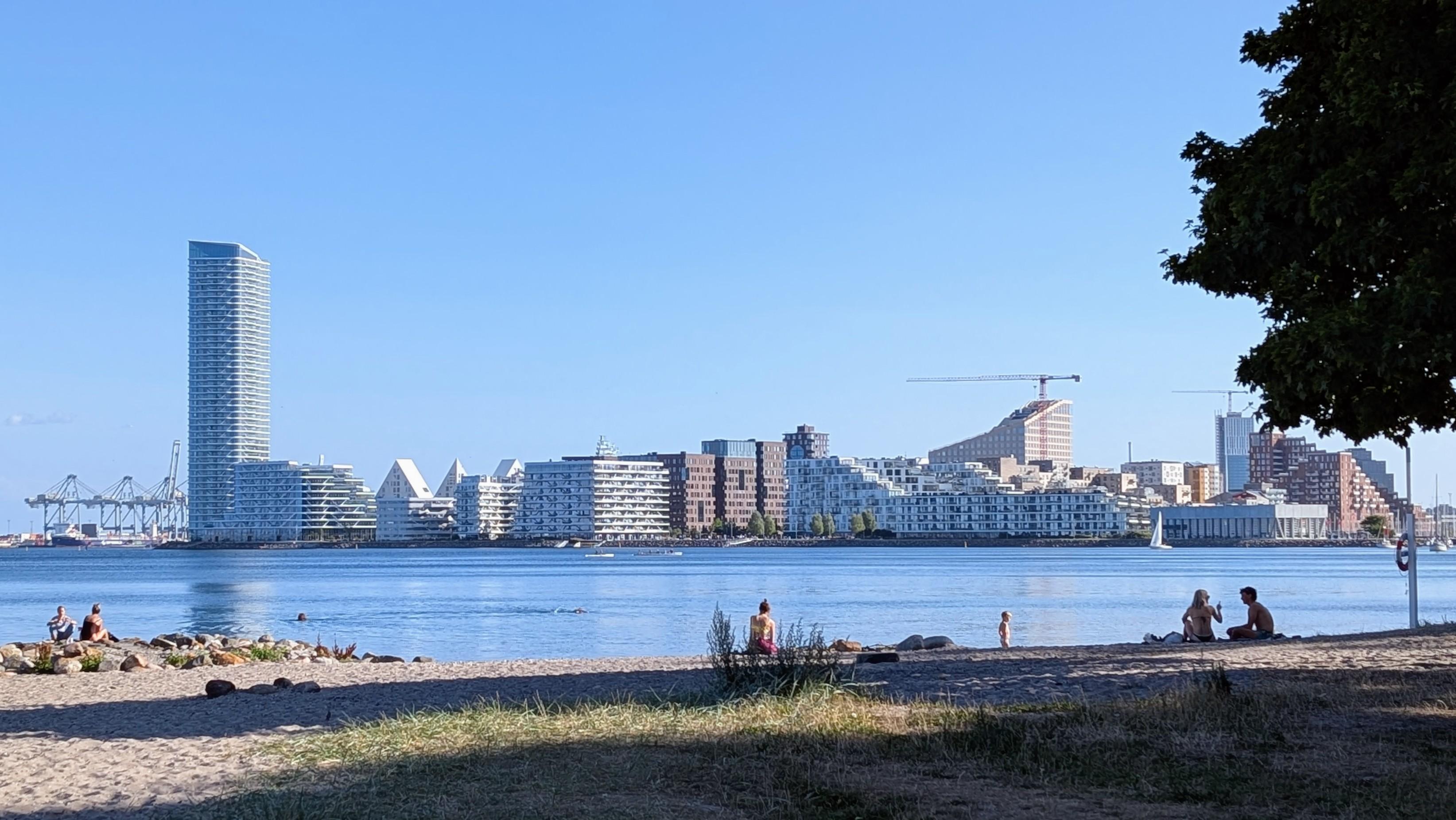 Modern skyline of Aarhus Ø, white is the dominating colour, a sandy beach with some people in the foreground, the sea dividing it from the town