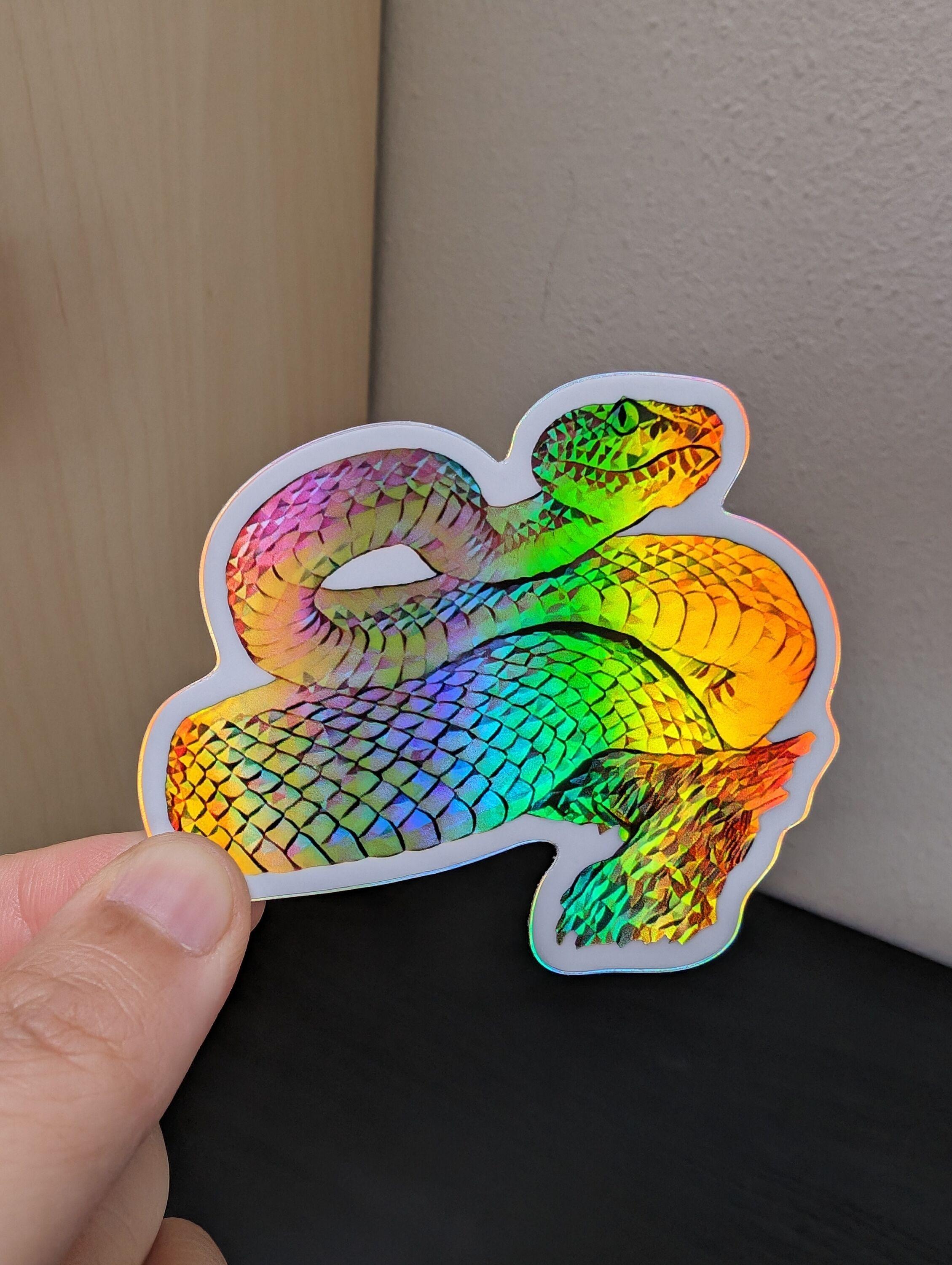 Photo of a left hand holding a diecut sticker. The sticker is a holographic snake drawing.