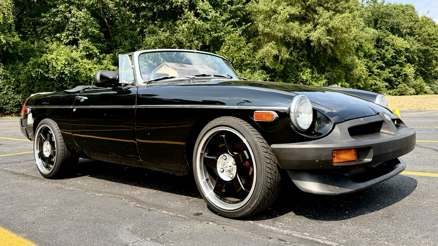 Photograph from a low angle at the front right of a black MGB with low profile tires in a parking lot with green shrubs in the background.