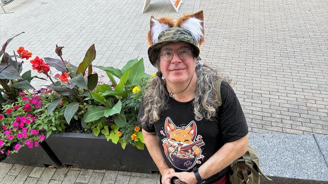 I, rather andogenous have long curly grey air bound up in low twintails routed under a hat and in front of my shoulders, adorned with fox ears, with a black t-shirt with a cartoon fox eating a bowl of ramen am looking up at camera on a selfie stick up in the air. to the left side of me find a planting of colorful flowers and behind a brick sidewalk