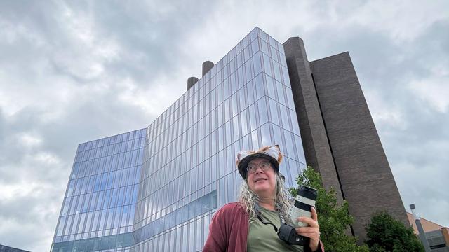 A glass, steel and brick tower rises behind a rather small looking man wearing fox ear hat, red hoodiie, green short and holding a camera with a big lens