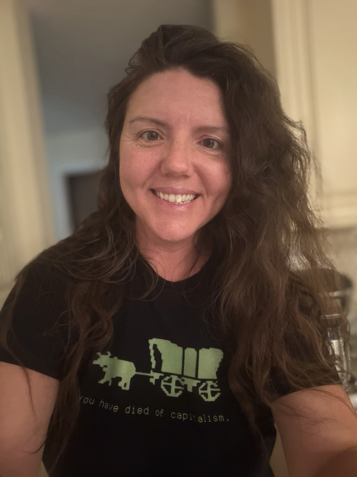Middle aged tired woman standing in kitchen wearing a black shirt with green Oregon trail logo (wagon pulled by ox) text underneath reads “you have died of capitalism”