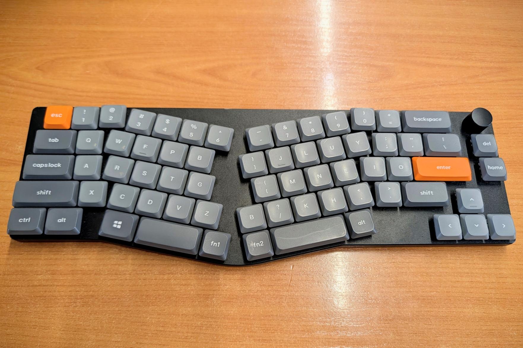 An ergonomic, split unibody, low-profile mechanical keyboard, in the Alice layout. The two halves angle inwards, with grey, orange-accented keycaps.