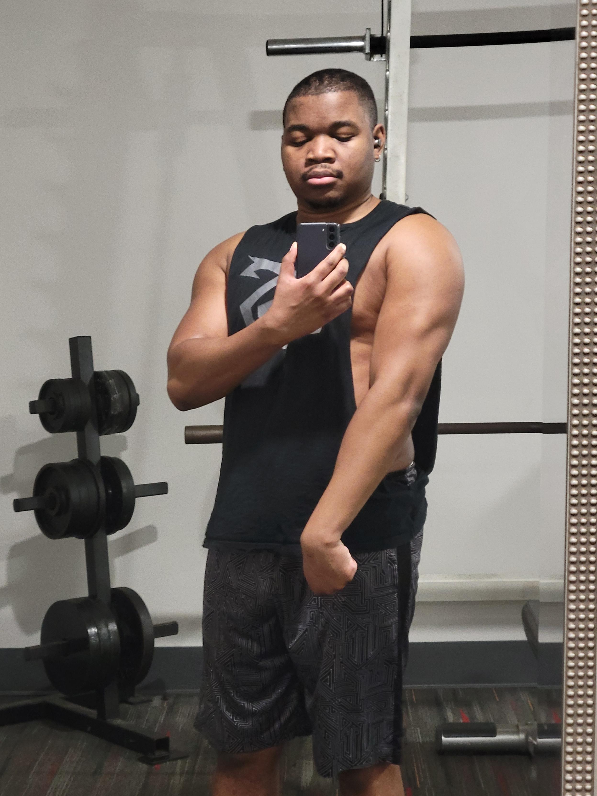 a mirror selfie of me wearing a sleeveless shirt and shorts in the gym and flexing my right tricep. there's plates and a squat rack in the background