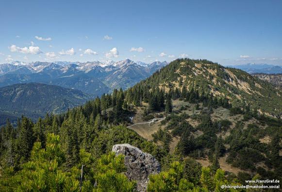 This image captures a breathtaking and expansive mountain landscape, showcasing the serene beauty of a high-altitude vista. The foreground features a rugged, rocky outcrop with patches of green vegetation, adding texture and a sense of wildness to the scene.
Beyond the outcrop, the landscape opens up to reveal a vast expanse of rolling hills and dense forests. The hills are covered in a rich tapestry of evergreen trees, their dark green foliage creating a vibrant and textured appearance. A wind…