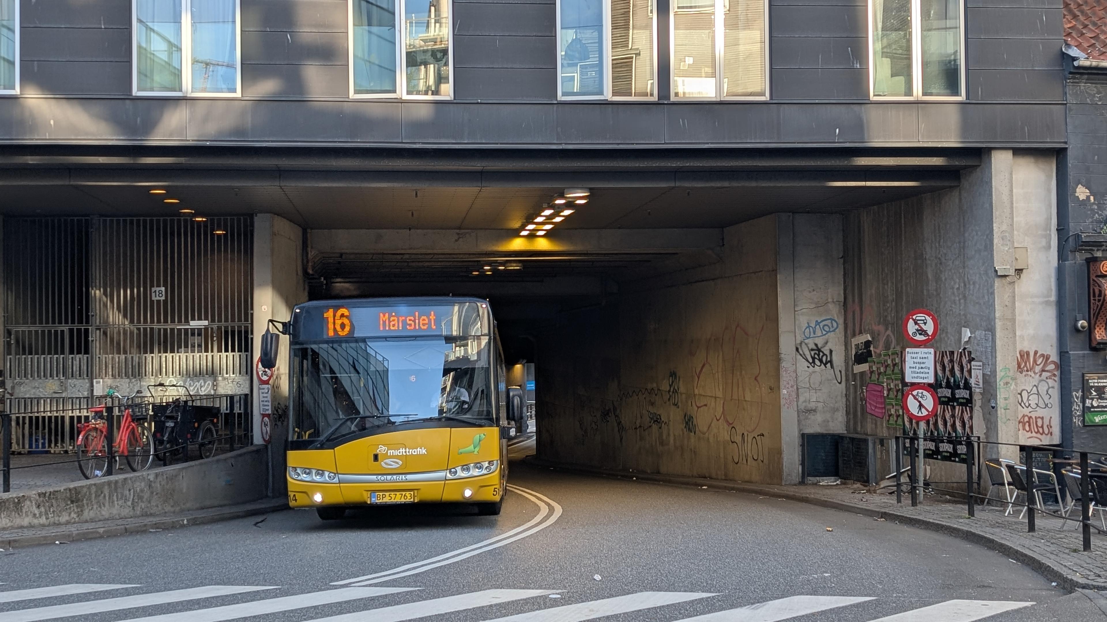 A two-lane passage leading through a building, a yellow bus as line 16 to Mårslet, a pedestrian crossing in front, bike parking on the left