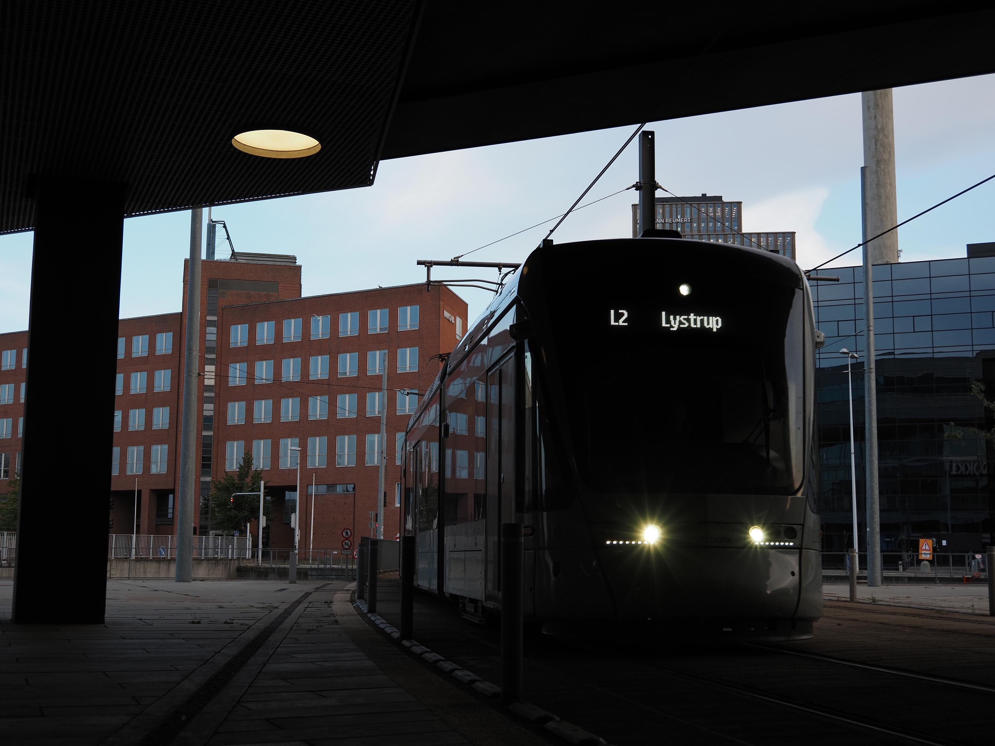 An oncoming tram as L2 to Lystrup underpasses a building, it appears almost fully dark with the headlights and destination indicator only, modern industrial buildings can be seen in the background