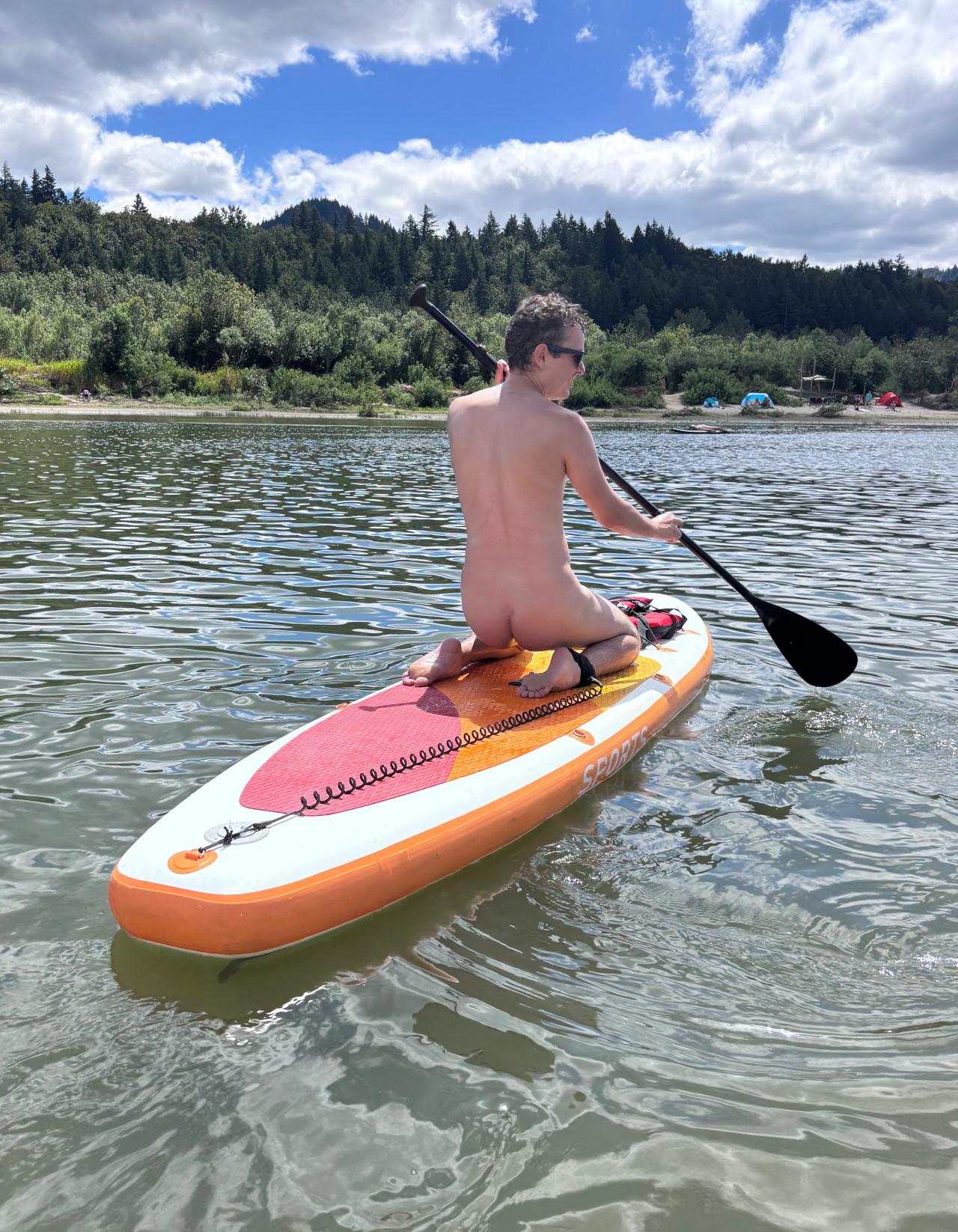A nude photo from behind of me sitting on my knees on the paddle board, with my full backside and bare bubble butt exposed.