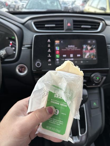 my hand holding a block of cheddar cheese. the wrapper has been haphazardly torn open. two large bites taken out of the top corners can be seen. the album thirteen tales from urban bohemia by the dandy warhols (2000) can be seen playing on the car's head unit